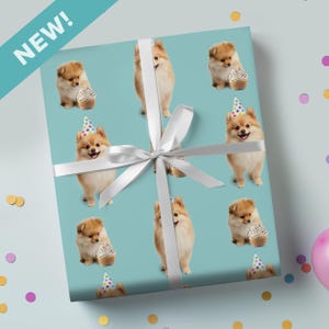 May include: Gift wrap with a pattern of Pomeranian puppies wearing party hats and holding cupcakes on a light blue background. The gift wrap is tied with a white ribbon. The word "NEW!" is in the upper left corner.