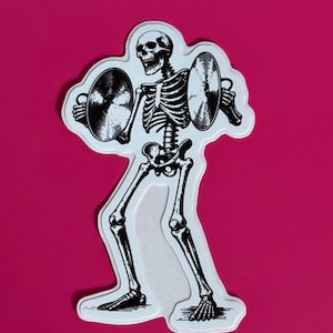 May include: A die-cut sticker featuring a black and white illustration of a skeleton playing cymbals. The skeleton is depicted in a dynamic pose, holding a metallic-looking cymbal in each hand, with detailed bone structures and shading throughout.