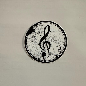 May include: A round sticker featuring a black treble clef symbol on a white background. The background has a distressed, speckled texture within a black border. The sticker is a music-themed item.