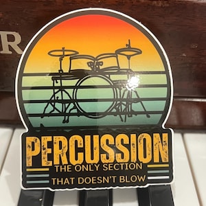 May include: A yellow and black sticker with a vintage sunset graphic and a drum set. The text reads "PERCUSSION THE ONLY SECTION THAT DOESN'T BLOW".