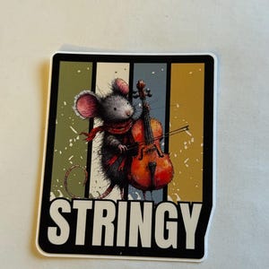 Op de afbeelding: Een sticker met een cartoonmuis die cello speelt. De muis, met een rode sjaal, staat tegen een achtergrond van groene, blauwe en gouden verticale strepen. Het woord "STRINGY" is in het wit gedrukt.