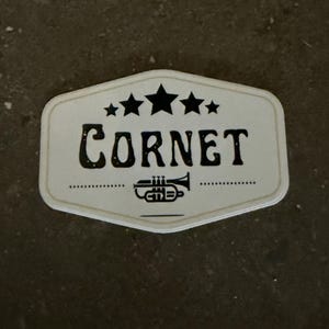 May include: A white, rectangular sign with rounded edges, featuring the word "CORNET" in bold, black letters. Above the word are three black stars, and below is a stylized cornet illustration. The sign has a vintage aesthetic.