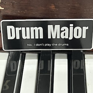 May include: Black and white sticker with the text "Drum Major" and the phrase "No. I don't play the drums".