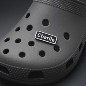 Puede incluir: Un zapato tipo zueco gris con múltiples agujeros redondos y una superficie texturizada. Un charm rectangular blanco y negro con el nombre "Charlie" está adherido al zapato.