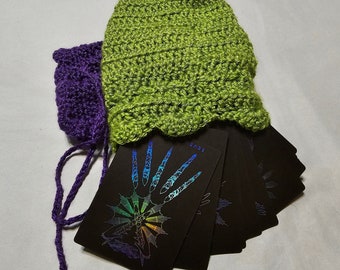 Puede incluir: Un juego de cartas de tarot negras con diseños de arcoíris iridiscentes, desplegadas y sostenidas en una bolsa de crochet verde y morada.