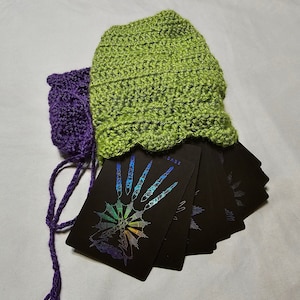 Puede incluir: Un juego de cartas de tarot negras con diseños de arcoíris iridiscentes, desplegadas y sostenidas en una bolsa de crochet verde y morada.