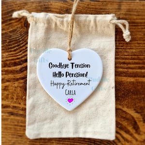 Puede incluir: Adorno de cerámica blanca en forma de corazón con el texto "Goodbye Tension Hello Pension! Happy Retirement CARLA" y un pequeño corazón rosa. El adorno está colgado de una cuerda de yute y está en una bolsa de tela blanca.