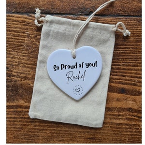 Puede incluir: Adorno de cerámica blanca en forma de corazón con el texto "So Proud of you! Rachel" y un pequeño diseño de corazón.