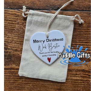 May include: White ceramic heart ornament with the text "Merry Christmas Work Bestie" and "Thank you for the laughs gossip & friendship" with a red and black plaid heart. The ornament is hanging from a string and is in a white drawstring bag. The text "Puddle Gifts" is in blue with a blue water drop graphic.