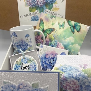 May include: A gift box containing a white mug with blue hydrangea prints, a card with butterflies and hydrangeas, a round ceramic ornament with the word love, a square tile with hydrangea art, and a towel featuring blue and purple hydrangea flowers.