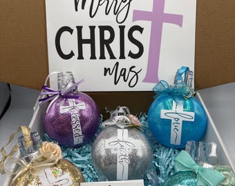 May include: A box set of Christmas ornaments. The set includes five glittery ornaments in various colors: gold, purple, silver, teal, and blue. Each ornament has a white cross design and a word like "blessed" or "believe". The box also contains a card that says "Merry Christmas".
