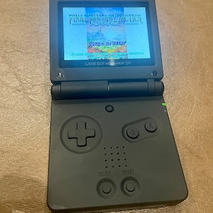 Peut inclure: Console Game Boy Advance SP noire, ouverte pour afficher le jeu "Final Fantasy Tactics Advance". L'écran affiche le titre du jeu et une invite à appuyer sur START. Les boutons et le pavé directionnel de la console sont visibles.