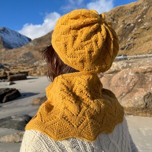 Puede incluir: Un gorro y una bufanda a juego de punto amarillo con un patrón de punto de cable. El gorro es de estilo boina y la bufanda es una capucha.