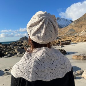 Puede incluir: Una persona lleva un gorro blanco de ganchillo y una chalina blanca de ganchillo con un patrón de encaje. La persona está de pie en una playa con una montaña en el fondo.