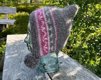 May include: A gray knit hat with a pointed top and a pom-pom. The hat features a pattern of pink and white stripes. The hat is displayed on a glass head form, set against a natural outdoor background.