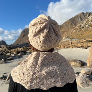 Puede incluir: Un gorro y una bufanda de punto beige con un delicado patrón de encaje. El gorro es de estilo boina con una corona redondeada. La bufanda es de estilo cuello alto con un borde festoneado. La persona lleva el gorro y la bufanda y está de pie en una playa con una montaña al fondo.