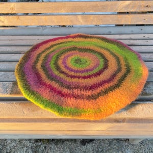May include: A round, colorful cushion with a spiral design. The cushion features concentric rings of orange, green, purple, and brown. The cushion is made of a soft, textured material and is resting on a wooden bench.
