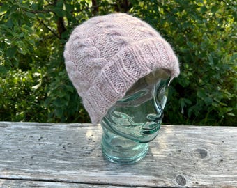 May include: A light pink knitted beanie with a cable knit design. The hat is made of soft yarn and is displayed on a glass head. The background features green foliage and a wooden surface.