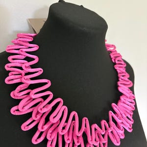 May include: A bright pink necklace featuring a layered, zigzag-patterned fabric design. The piece consists of multiple rows of soft, tubular material arranged in a continuous, undulating wave formation that encircles the neck area.