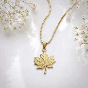 Peut inclure: Un collier doré avec un pendentif en forme de feuille d'érable détaillée suspendu à une chaîne fine. Le pendentif présente une surface texturée qui imite les nervures et les bords naturels d'une feuille, créant un aspect botanique réaliste.