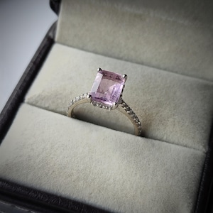 May include: A silver ring featuring a large, rectangular, pale purple gemstone. The band is embellished with small, clear stones. The ring is presented in a black jewellery box with a grey interior, offering an elegant display.