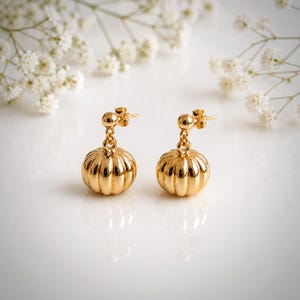 May include: A pair of gold-toned drop earrings featuring a small, polished sphere attached to a larger, ribbed, pumpkin-shaped charm. The earrings have a reflective, metallic finish and are designed with a post-back closure for secure wear.