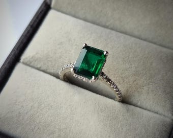 May include: A silver ring with a large, emerald-cut, deep green gemstone. The band is encrusted with small, clear stones. The ring is presented in a black jewelry box with a gray interior.