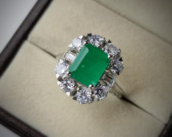 May include: A silver ring featuring a large, rectangular emerald-cut green gemstone, surrounded by a halo of smaller, round, clear gemstones. The ring is set in a silver band and is displayed in a brown jewelry box.
