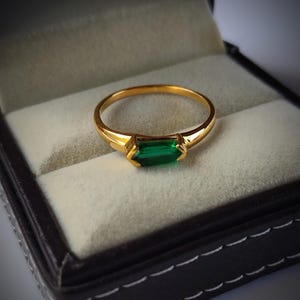 May include: A gold ring with a rectangular, emerald-green gemstone. The ring is set in a gold band and is displayed in a black jewelry box with a light gray interior. The ring is a classic piece of jewelry.