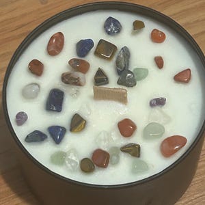 May include: A white candle with a variety of small, colorful stones embedded in the wax. The stones are in shades of red, blue, green, brown, and white.