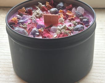 May include: A black candle jar with a pink wax top. The wax is filled with small purple and pink crystals, dried rose petals, and silver sprigs.