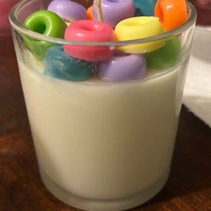 Puede incluir: Un tarro de vidrio transparente lleno de cera blanca y cubierto con velas de colores en forma de donut. Las velas son de color verde, morado, amarillo, naranja y rosa.
