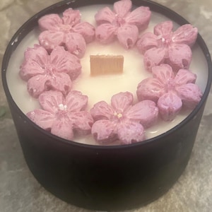 May include: A black round candle with a cream-colored wax interior. The candle is decorated with a ring of pink flower-shaped embellishments. A wooden wick is centered in the candle.