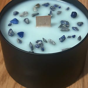 May include: A black candle with a light blue wax top. The wax is embedded with small blue and gray stones. A wooden wick sits in the center of the candle.