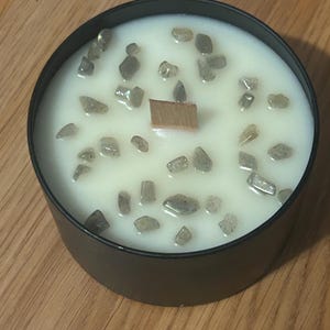 May include: A black candle jar with a white wax base and a wooden wick. The wax is embedded with small, gray, iridescent stones.