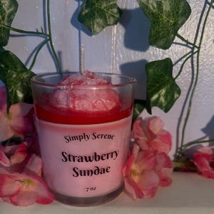 May include: A clear glass candle with a pink wax base and a textured pink wax topping resembling a sundae. The label reads Simply Serene Strawberry Sundae 7 oz in black text. A single white wick extends from the center of the wax topping.