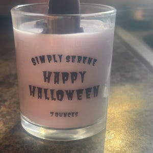 May include: A clear glass candle container filled with light purple wax, featuring a dark purple crystal-like topper. The front displays black text reading SIMPLY SERENE HAPPY HALLOWEEN and 7 OUNCES in a stylized, jagged font.
