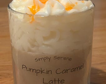May include: A clear glass candle with a layered design. The bottom layer is a light brown color, with the words "Pumpkin Caramel Latte" printed on it. The top layer is white with orange accents, resembling whipped cream. The candle is 10 oz soy wax.