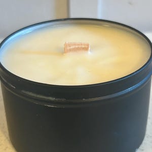 以下が含まれることがあります： 黒いキャンドルジャーに木製の芯と薄い黄色のワックスが入っています。ジャーはワックスで満たされ、芯はジャーの中心に置かれています。