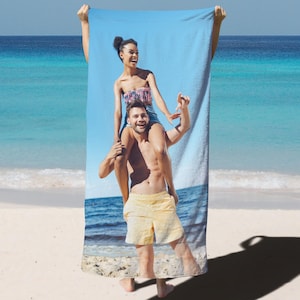 May include: A beach towel featuring a couple on a sunny beach. The towel displays a vibrant image of a woman sitting on a man's shoulders, with the ocean in the background. The towel is held up against a backdrop of the sea and sky.