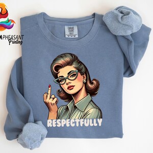 May include: A blue long-sleeve sweatshirt featuring a retro-style illustration of a woman wearing cat-eye glasses and a button-up shirt, giving the middle finger. The word "RESPECTFULLY" is printed below the illustration.