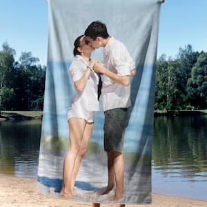 May include: A beach towel featuring a couple kissing on a beach, with a blue sky and water background. The couple is wearing white shirts and shorts. The towel is held up by unseen hands.