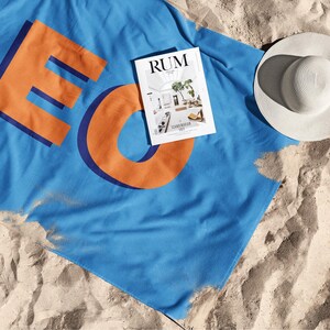May include: A blue beach towel with the word "LEO" in orange letters and a magazine titled "RUM" on top of it. A white sun hat is also on the sand. The items are arranged on a sandy beach.
