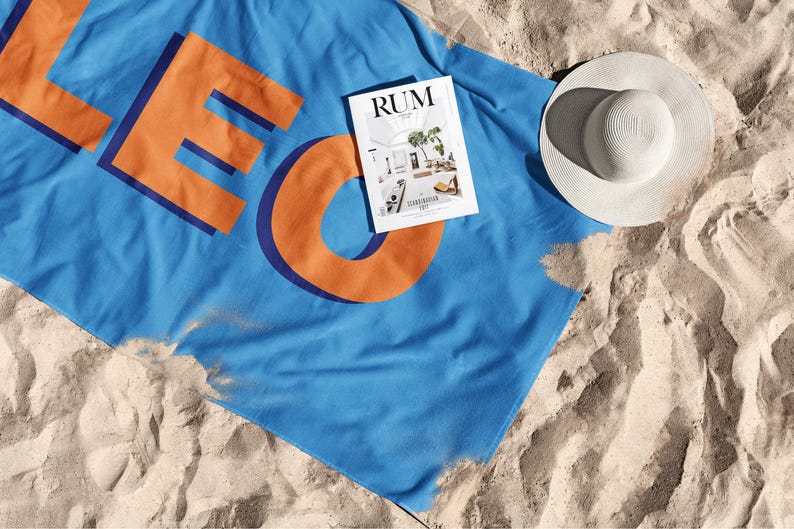 May include: A blue beach towel with the word "LEO" in orange letters and a magazine titled "RUM" on top of it. A white sun hat is also on the sand. The items are arranged on a sandy beach.