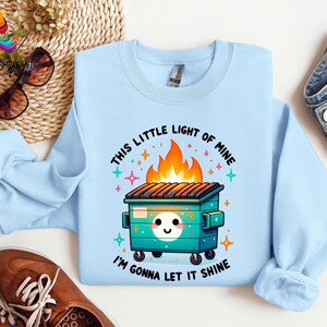 Pu&ograve; includere: Felpa blu chiaro con un'illustrazione di un cassonetto della spazzatura in fiamme in stile cartone animato. Il testo recita "This little light of mine, I'm gonna let it shine."