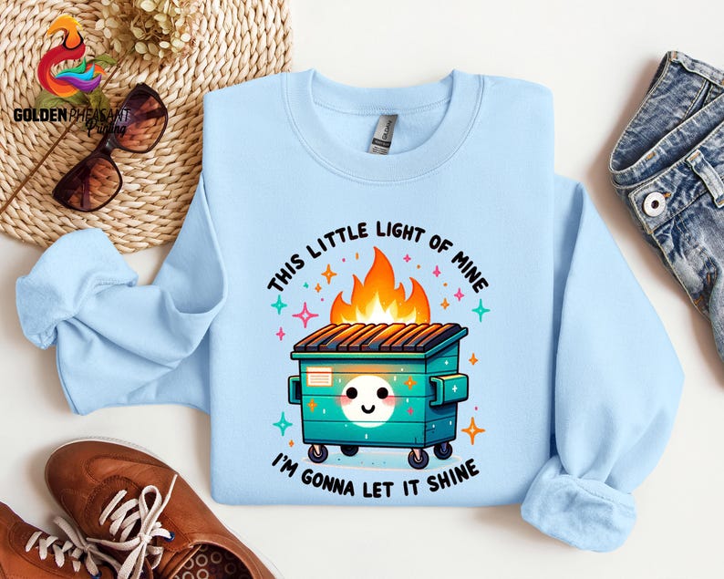 Pu&ograve; includere: Felpa blu chiaro con un'illustrazione di un cassonetto della spazzatura in fiamme in stile cartone animato. Il testo recita "This little light of mine, I'm gonna let it shine."