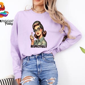 May include: A light purple long-sleeve sweatshirt featuring a retro illustration of a woman wearing glasses and giving the middle finger. The word "RESPECTFULLY" is printed below the illustration. The sweatshirt is paired with blue jeans.
