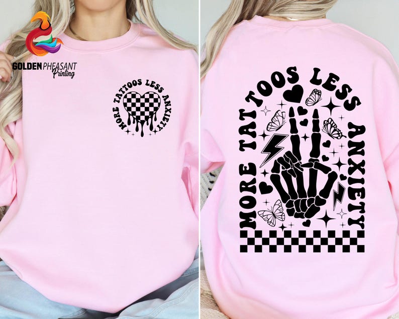 Pu&ograve; includere: Felpa rosa con testo nero che recita "Pi&ugrave; tatuaggi, meno ansia" con un cuore a scacchi bianco e nero, una mano scheletrica e altri elementi grafici.