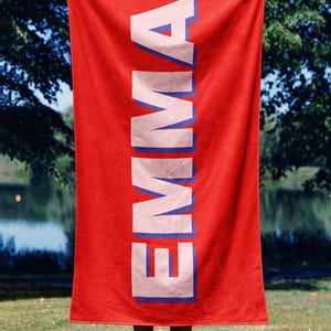 May include: A red rectangular beach towel with the name "EMMA" printed vertically in large, white capital letters with a blue outline. The towel is held up against a background of trees and a body of water.
