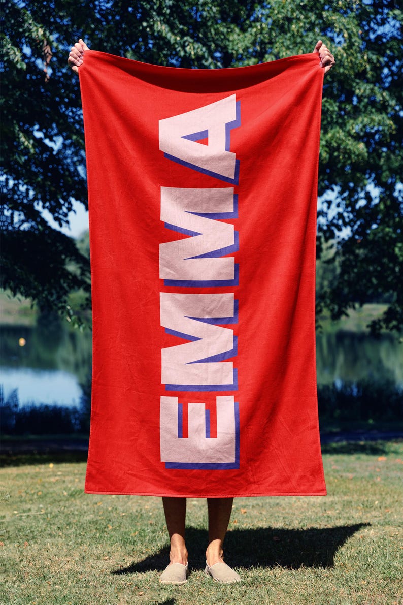 May include: A red rectangular beach towel with the name "EMMA" printed vertically in large, white capital letters with a blue outline. The towel is held up against a background of trees and a body of water.
